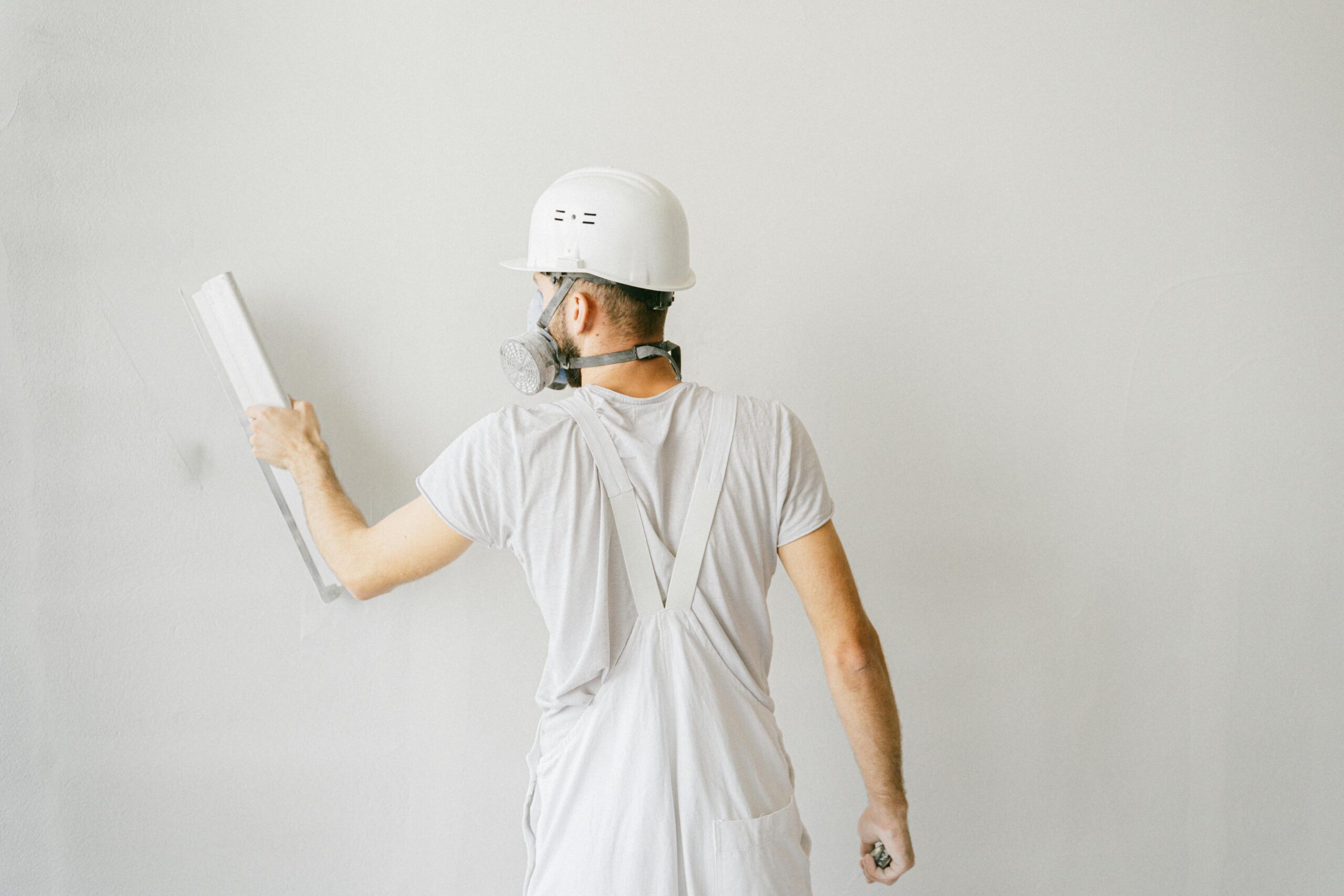 Servicios Back view of a construction worker in safety gear plastering a white wall.